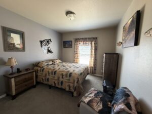 Guest bedroom showcasing a full bed with brown bear and moose-patterned quilt and pillows. Light gray walls adorned with metal jumping salmon art piece, two framed landscape photos, wood dresser, lamp and clock on a nightstand, gray recliner with blanket, flush ceiling light, plaid curtains over bright windows, and beige carpet.