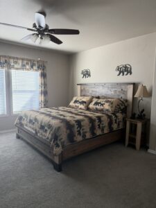 Cozy master bedroom with a rustic wooden king bed frame covered in a black bear-patterned quilt and pillows. Tan walls feature two metal bear silhouette decorations, a ceiling fan, lamp on wood nightstand, and plaid curtains over sunny windows; beige carpet flooring completes the space.