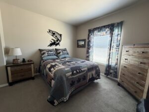 Guest bedroom with a full wood bed frame draped in a blue-toned quilt featuring maple leaves and black bears. Tan walls display a large metal salmon silhouette art piece and landscape painting; includes wood dresser, lamp, nightstand with clock, plaid curtains over sunny windows, and beige carpet floor.