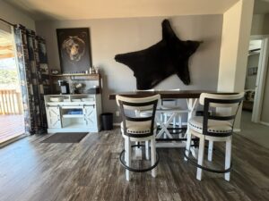 Rustic dining area with white farmhouse table and four black leather upholstered bar stools on wood flooring. White cabinet with blue shutter doors holds coffee maker and storage; gray walls feature two large black bear art pieces; plaid curtains frame open door to deck overlooking outdoors.