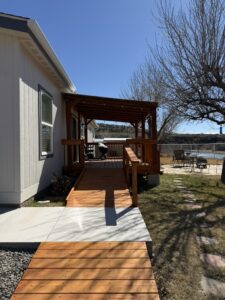 Exterior view of a light blue house with white trim and wooden accents in a desert landscape, featuring an orange wood ramp leading to a covered porch with railing. A charcoal grill sits on the porch near Adirondack chairs and a small table overlooking a calm river, with bare trees, grass, gravel path, and distant rocky cliffs under a clear blue sky.